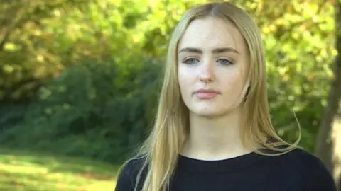 A young woman with long blonde hair, wearing a black top, looks at the camera.