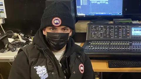 BBC JM in the music studio, he's in a body warmer with a sticker on the breast which reads RIP Harvey and Kyres. Forever Young. He's wearing a wooly hat and a black face mask.