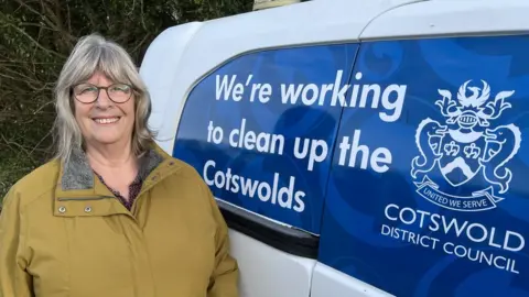 Ms Layton wears glasses and a mustard yellow coat, looking into the camera next to a council-branded van