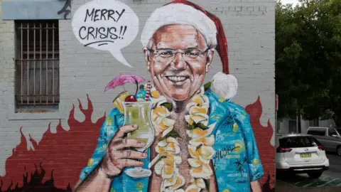 Getty Images A wall mural depicting Scott Morrison, dressed in a Hawaiian shirt and Santa hat while holding a cocktail against a backdrop of flames, saying "Merry Crisis".