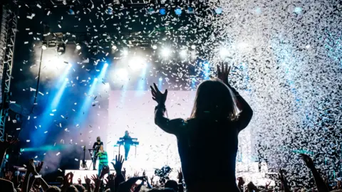 A girl enjoying a band playing at the Big Feastival as she sits on someone's shoulders and confetti falls