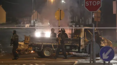 PA Police in riot gear patrol past a burnt-out vehicle during the violence in Londonderry