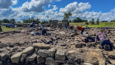 Vindolanda Trust A group of people on their knees digging between rocks and stones at the Roman settlement