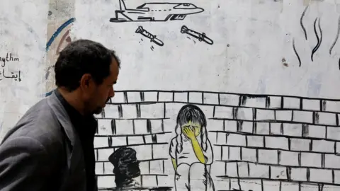 EPA A man walks past a wall covered in graffiti depicting a warplane dropping bombs on child, in Sanaa, Yemen (10 August 2018)