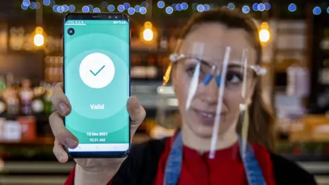 PA Media A woman working in a bar holds a phone displaying an approved Covid certificate