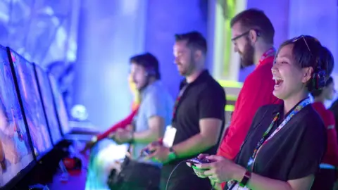 Getty Images Four players at four gaming stations hold controllers, with the closest person laughing out loud at the unseen events on her screen