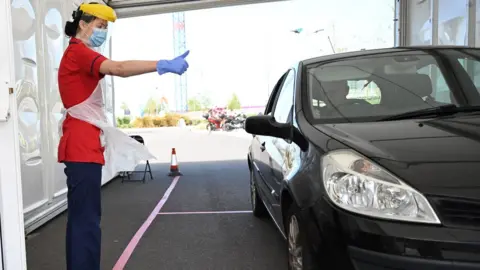 Getty Images A member of clinical staff wears Personal Protective Equipment (PPE) as she gestures to a key worker at a drive-through test centre for the novel coronavirus at Royal Papworth Hospital in Cambridge on May 5, 2020. - NHS services have come under increased strain with the number of a patients hospitalised and requiring critical care because of the COVID-19 pandemic which has claimed over 30,000 lives in the UK. Mass testing has become a key part of the UK strategy in their battle against the virus.