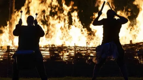 PA Media Members of the Pentacle Drummers drum in front of a burning ceremonial Viking long boat at Butser Ancient Farm