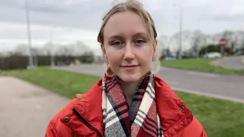 Claudia Bradley with straight blonde hair wearing a red top outside