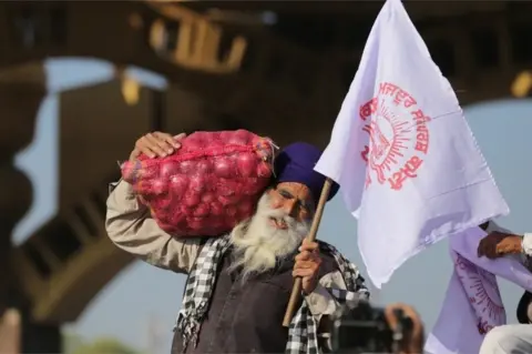 EPA A farmer carries a bag of onions and a flag prior to the start of a march towards New Delhi to protest against new farm laws, on the outskirts of Amritsar, India, 27 November 2020.
