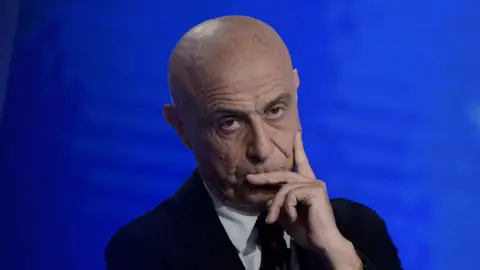 Getty Images Marco Minniti is pictured against a bright blue background, one hand against his face in a pensive pose