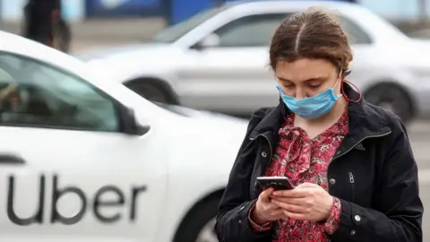 Getty Images A woman wearing a face mask uses a phone as an Uber passes by in the background.