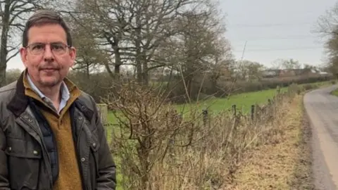 Mr Mitchell stands to the left of the picture looking into the camera in front of a country lane and narrow field