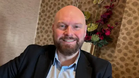 David Jones, a shaven-headed white man in a suit smiles in the camera.