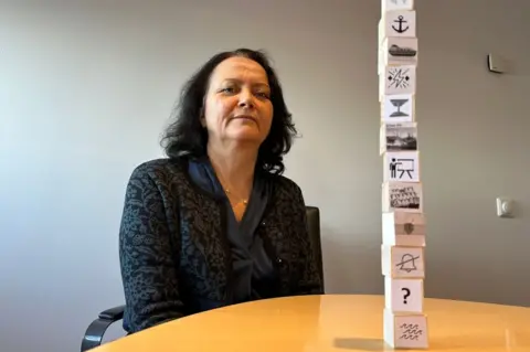 BBC A woman sits next to a table on which are stacked 12 small wooden bricks each with a different image drawn on