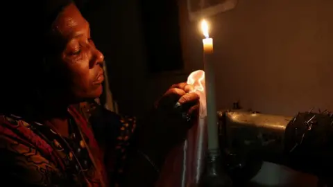 Reuters Dressmaker Faieza Caswell from Mitchells Plain sews under candlelight in her workplace, on the Cape Flats due to South Africa's struggling power utility company Eskom, implementing regular power cuts - called 'load-shedding', in Cape Town, South Africa February 11, 2023