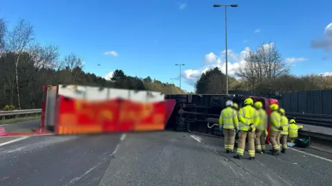 X/National Highways East Red lorry overturned on the M40 and people in high-vis jackets standing near it