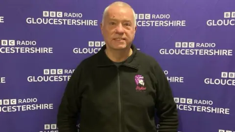 BBC Nick Gazzard stood smiling in front of BBC Radio Gloucestershire backdrop