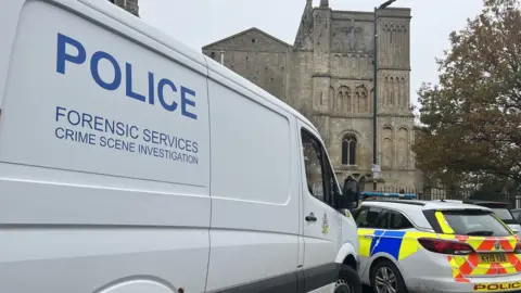 The side of a police forensics van, part of a police car and the abbey in the background.