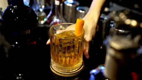 A person puts whiskey in a tumbler in a bar along with drink bottles.