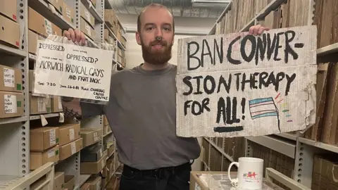 Adam Baker Adam Baker at Norfolk Heritage Centre holding up a poster and a sign that are in the Queer Norfolk archive