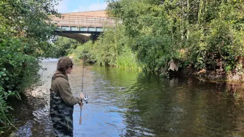 PA Media An angler holding a fishing rod and wearing waders stands in the River Tone