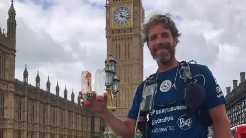 Handout Mr Tinga holding up plastic bottles in front of Big Ben in London.