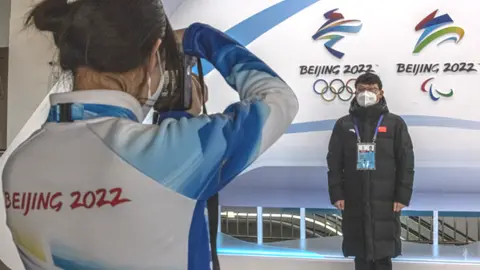 Reuters A woman snaps a photo of a man wearing a face mask next to the logos of the Beijing 2022 Olympic and Paralympic Winter Games inside the Olympic main media centre