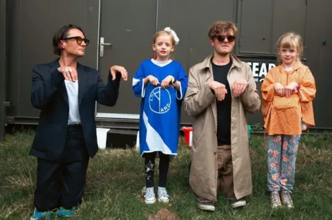 Jamie Macmillan Ryan Needham and James Smith of Yard Act with two children at Latitude Festival, pretending to be meerkats