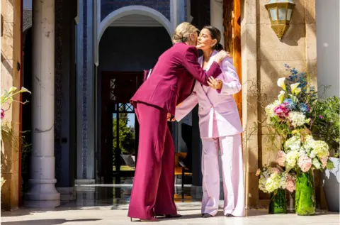 Getty Images Queen Maxima of The Netherlands visiting Princess Lalla Meryem of Morocco. The two women are wearing purple suits of different shades and are pecking each other on the cheek.