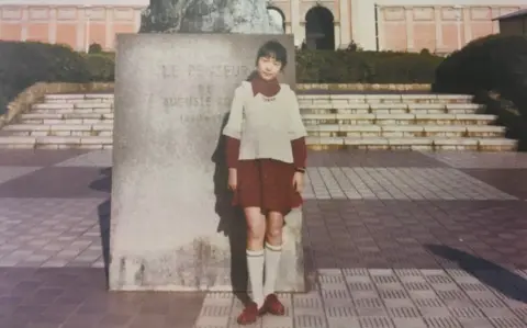 Yokota family Megumi stands for a picture in a red dress, white jumper and long knee socks