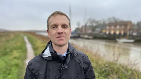 Shaun Whitmore/BBC Man with short light-brown hair wearing a blue raincoat stands alongside the estuary