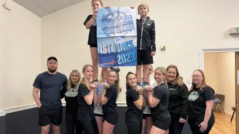 BBC The cheerleaders pose to the camera with a big sign saying 'Winter weekender 1st place 2023'