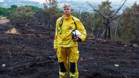 BBC/Claire Marshall Dave Swallow standing in a yellow outfit against charred moorland.