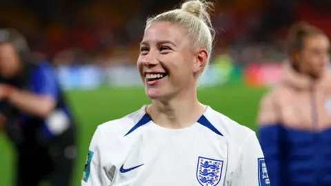 Getty Images Bethany England of England during the FIFA Women's World Cup Australia & New Zealand 2023 Round of 16 match between England and Nigeria at Brisbane Stadium. Bethany is a 29-year-old white woman with long blonde hair tied in a bun at the top of her head. She wears the white England shirt complete with three lions logo and blue trim. She smiles into the crowd.