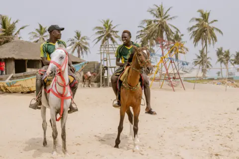 Grace Ekpu/BBC Two men riding horses on the beach