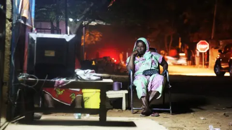 Fatoumata Diabate Woman sits in chair outside watching TV