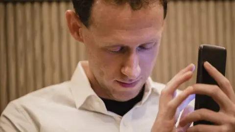 SolStock/Getty Images A close-up of a man wearing casual clothing, he has his smartphone in his hand and he is using a visually impaired mobile app to help assist him