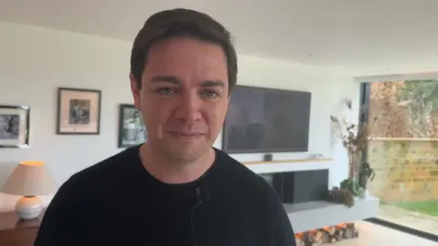 Duncan Greenaway with short dark hair and blue sweater standing in modern living room