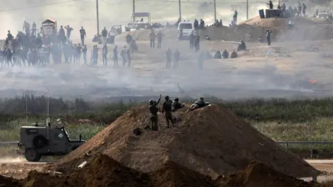 Reuters Israeli soldiers stand on a berm near the Israel-Gaza border fence, as Palestinians protest on the other side (6 April 2018)