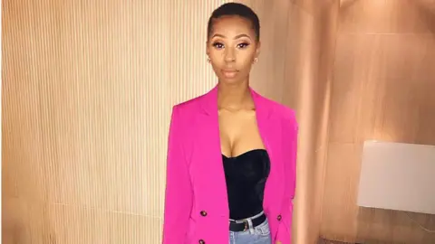 Photo From Luan Luan is standing in front of a wood panelled background wearing a bright pink jack a black top and jeans