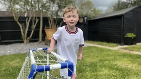 Lauren Carter/BBC Jesse standing in his garden holding the crossbar of a small football goal