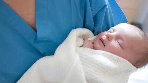 Getty Images/IAN HOOTON Newborn baby held by maternity unit worker