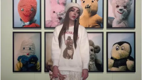 Kellen Beckett Kellen is standing in front of eight framed pictures of different animal teddy bears. She is wearing a white knitted jumper that features a picture of a brown rabbit with a pink bow tied around its ears. She has long brown straight hair and is wearing a white beret with a small group of pearls on the left hand side of it. Kellen is posing side on and is looking down towards the floor rather than towards the camera.
