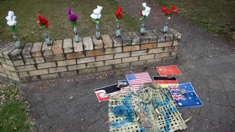 Getty Images A small tribute memorial in the New Zealand town of Whakatane shows flowers and the flags of countries where victims were from