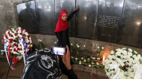 EPA A Kenyan woman (C), who lost her relative, poses for a photo being taken by her friend as she stands in front of the plaque bearing the names of those killed during the 1998 bombing of US Embassy, at the August 7th memorial park in Nairobi, Kenya, 07 August 2018.