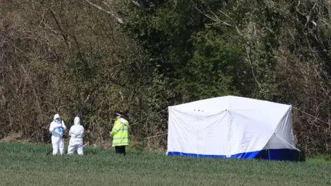 PA Media Police erected a white tent on the edge of a field at Akholt Wood