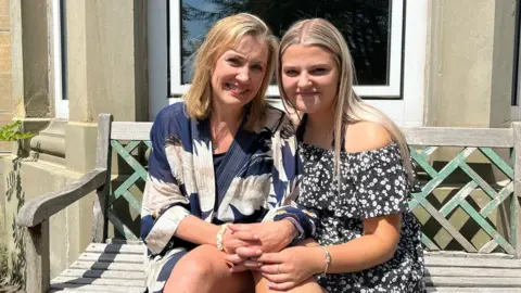 Nicola and Gracie Nuttall sitting together on a bench in their garden. Nicola is on the left and is wearing a blue and white dress as she sits with her hands clasped on her lap. Gracie is to her right and has her hand on her knee, she's wearing a black and white floral dress