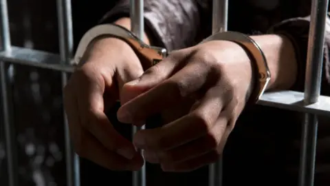 Getty Images A prisoner behind bars with hands cuffed