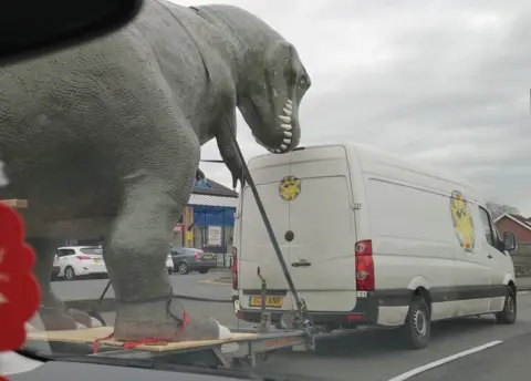 The dinosaur on the back of the trailer on the road